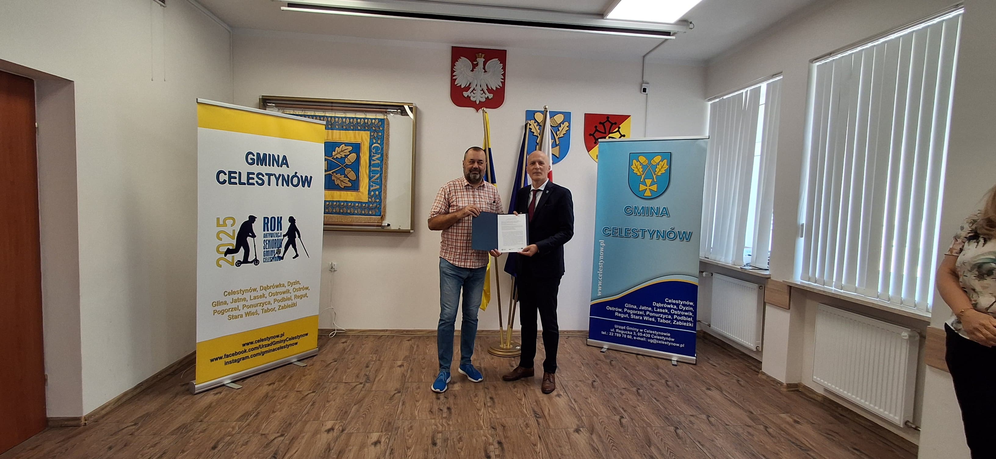 dwie osby stojąca na tle roll-upów i przekazujące sobie umowę